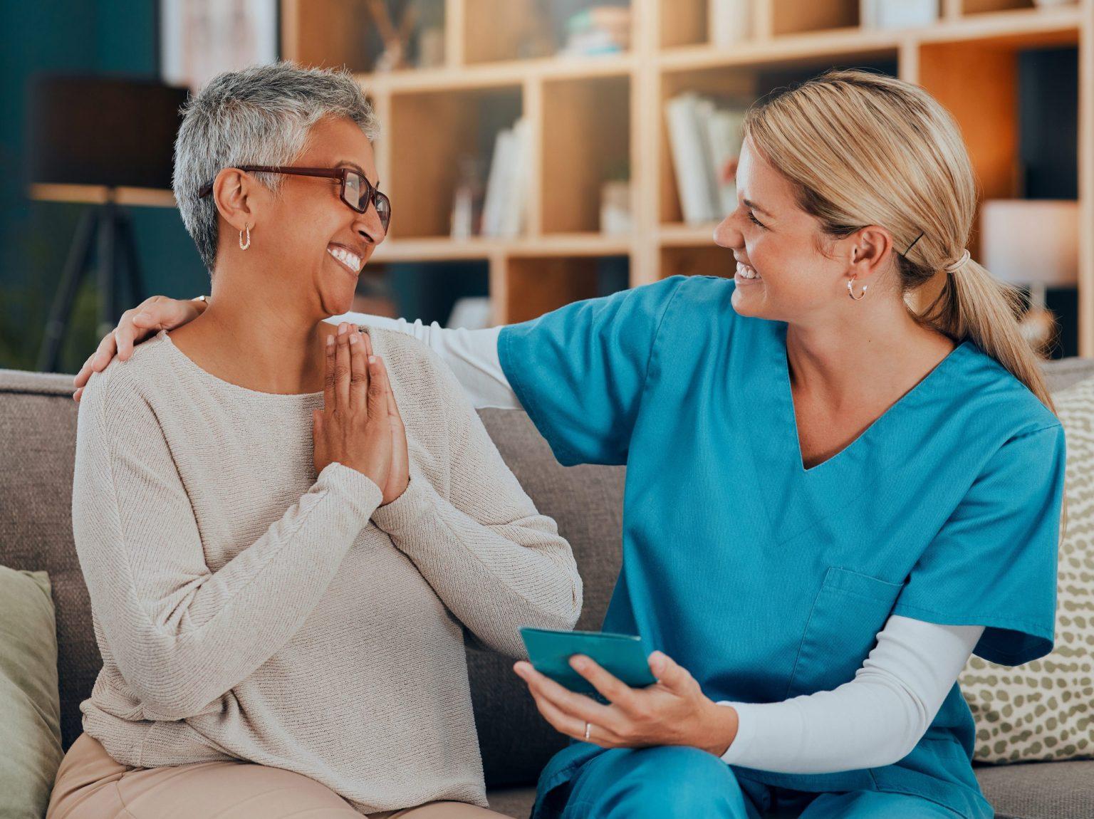 Photo of a female care provider with an older woman, smiling together on a couch.