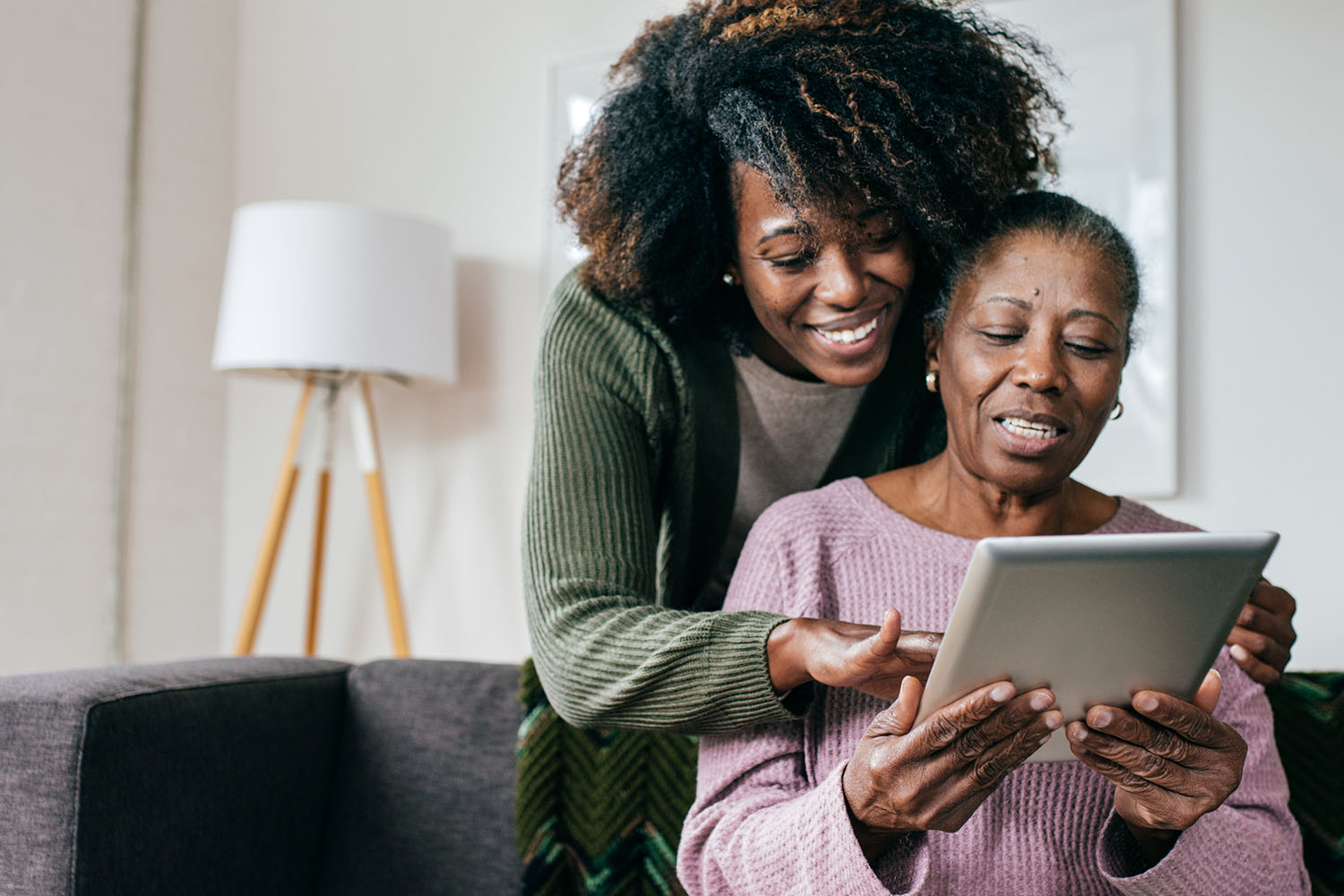 Photo of an adult daughter looking over the shoulder of her older mother and assisting with a digital tablet.