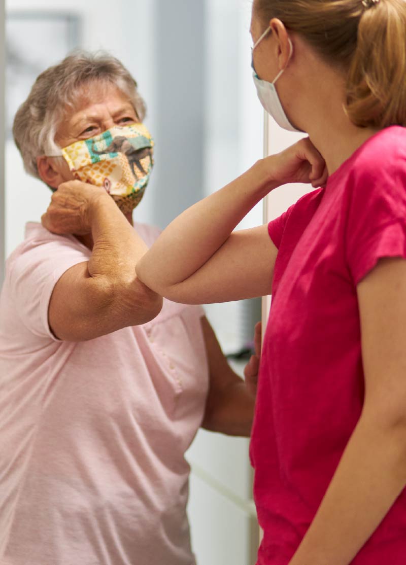 Photo of a female care provider giving an elbow-bump to an older woman, both wearing protective masks.
